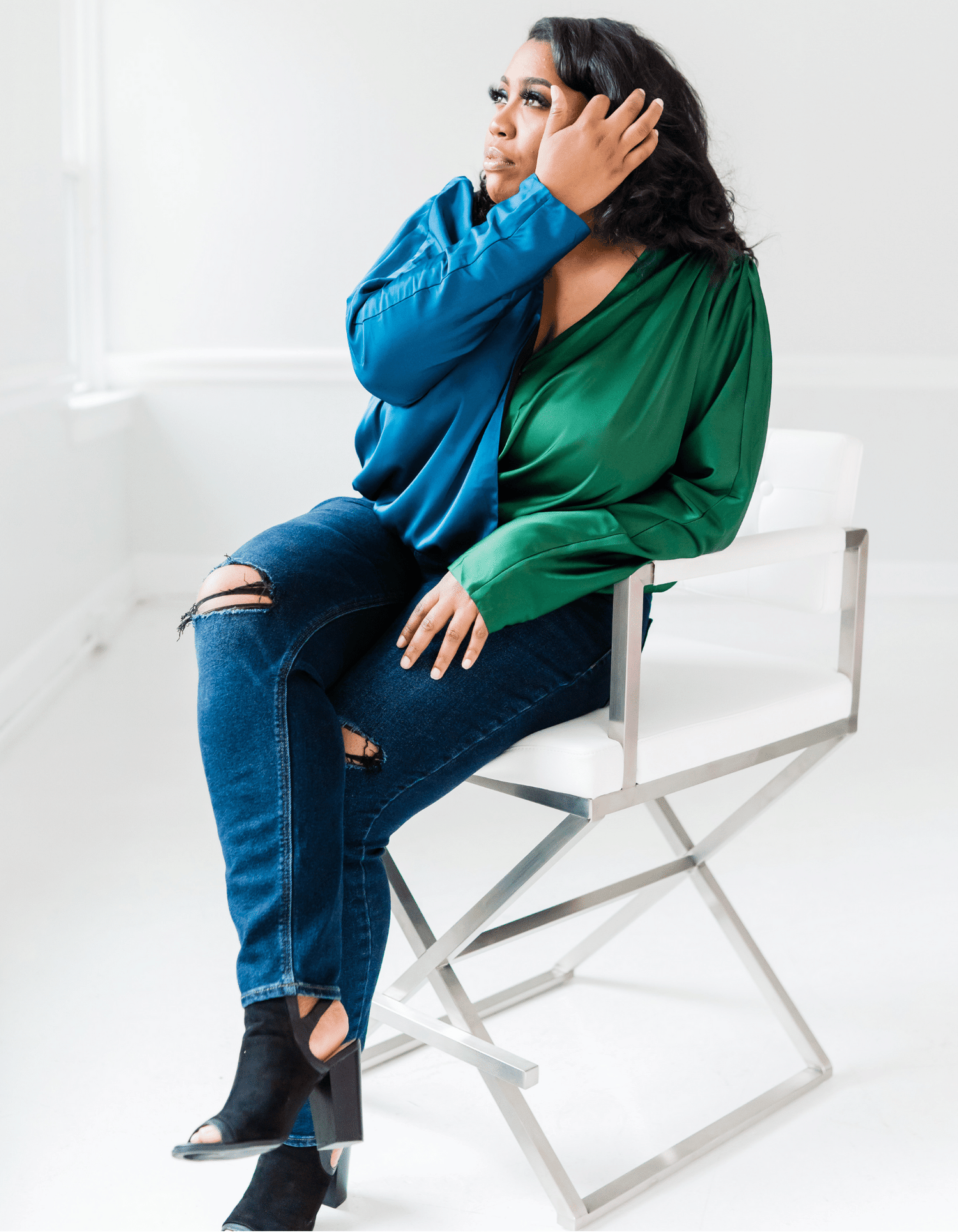 A black woman sits in a tall chair, her legs are crossed while her hand gently moves her soft black curls out of her face. The room is all white and full of natural light.